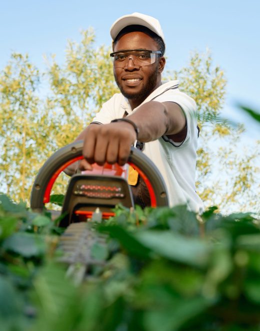 Portrait of cheerful african man in protective glasses and summer hat pruning hedge with hand trimmer. Male gardener in working clothing cutting bushes during sunny days.